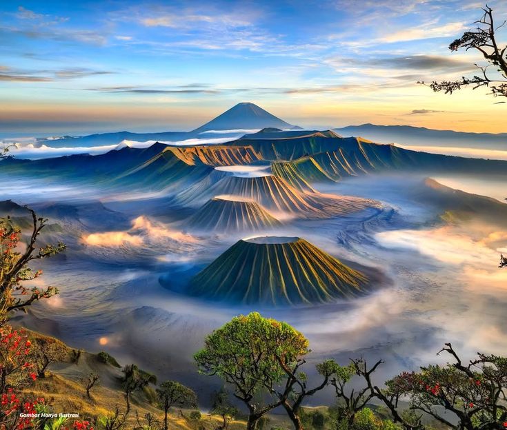 A breathtaking view of Mount Bromo in East Java, Indonesia, with its iconic smoking crater surrounded by a sea of mist and dramatic volcanic landscapes. The scene is framed by the majestic Mount Semeru standing tall in the background under a clear blue sky. The golden morning light enhances the rugged beauty of the mountains, creating one of the most unforgettable sunrise views in the world. This natural wonder is a must-visit destination for travelers, photographers, and adventure seekers exploring Indonesia.